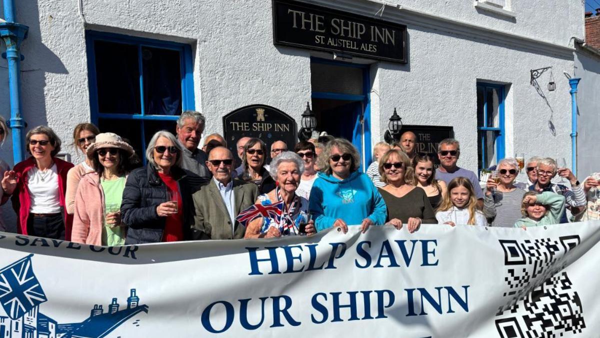 A group of people stand in front of a pub holding glasses and a banner which reads 'Help Save Our Ship Inn'. 