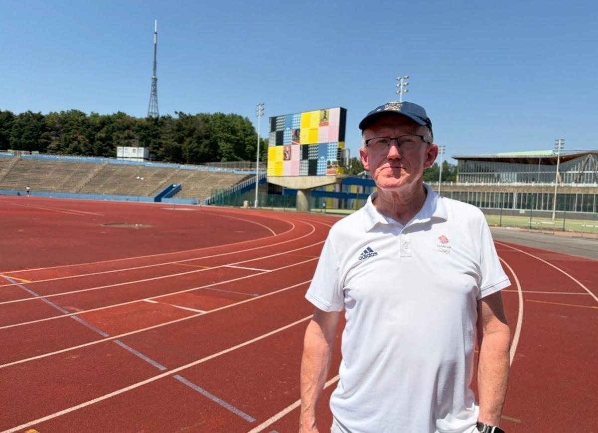 A man in a white Team GB T-shirt stands on Crystal Palace track with the iconic scoreboard in the background covered by netting.