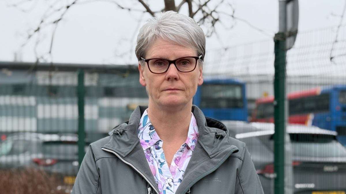 A woman with white hair, and wearing black-rimmed glasses, and a grey jacket over a floral shirt.