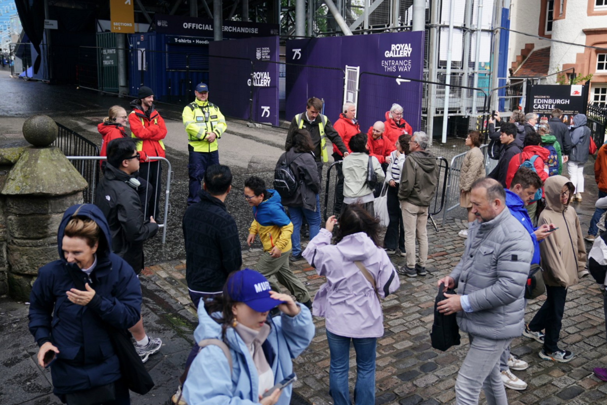 A crowd of about 20 people on a windy cobbled street, a woman holds down her hat while another woman has her hood blown from her head. Staff in high vis vests stand at a metal barrier.