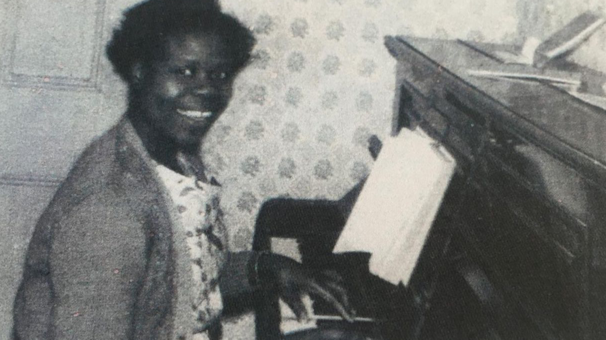 Grace Adebowale is pictured in her 40s in a black and white photo. She is smiling at the camera while seated at a piano which she appears to be playing.