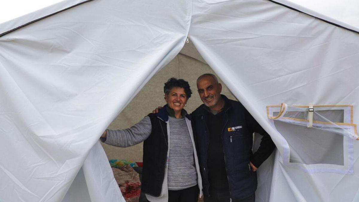 A man and a woman stand in the entrance of a large white tent. They are smiling and the man has his arm around the woman. 