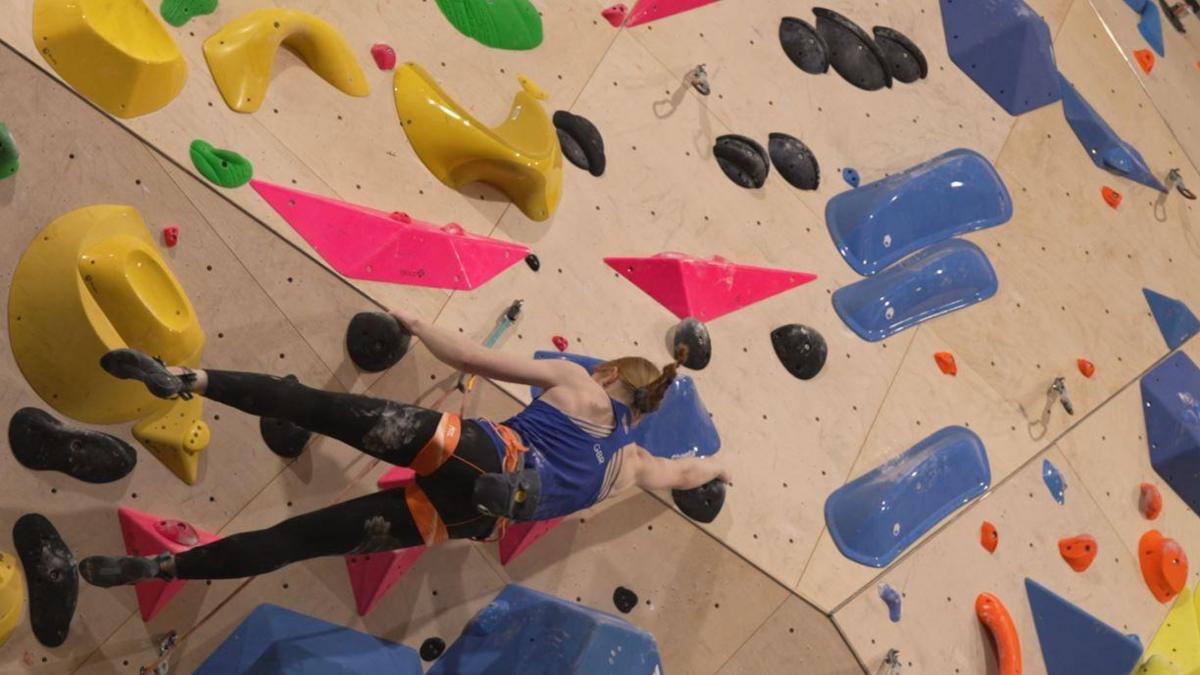 Connie Bridgens, wearing black leggings and a blue top, climbing up a multi-coloured wall. 