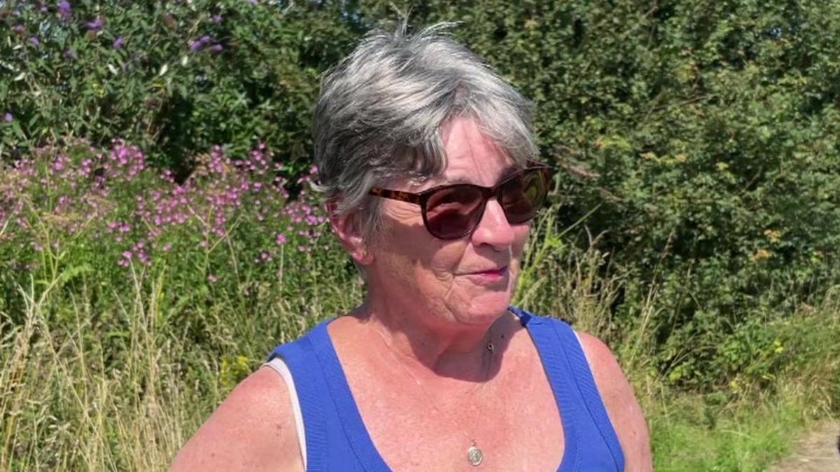 Head shot of Sherford resident Fran Cook stood in front of a grass verge with pink flowers growing in the background.