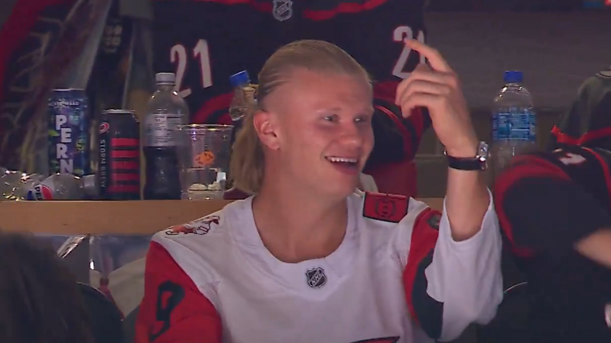 Erling Haaland, wearing a white Carolina Hurricanes jersey with red, black and white sleeves, smiles and points his left hand in the air