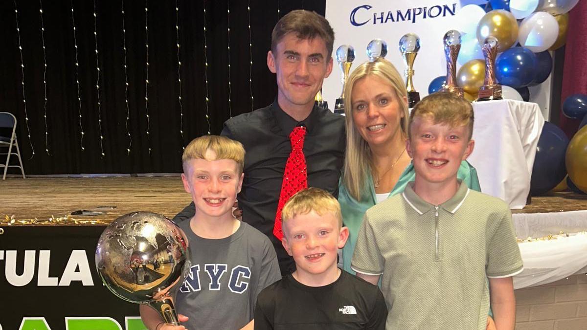 A man, with short brown hair, wearing a black shirt and red tie. He is standing beside a woman with blonde hair and three young boys, all with blonde hair.