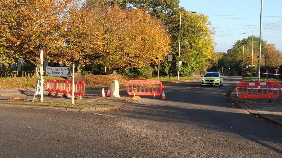 A road closed in Bedford. There is one road sign, a police car, and red bollards and cones. There are a number of autumnal trees and lamp posts. 