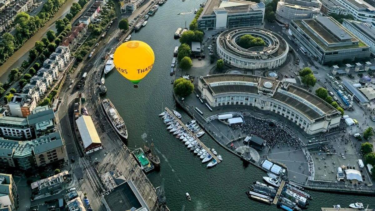 A yellow Thatchers hot air balloon hovers over Bristol's harbourside with the Lloyd's Ampitheatre and boats visible below, along with the River Avon