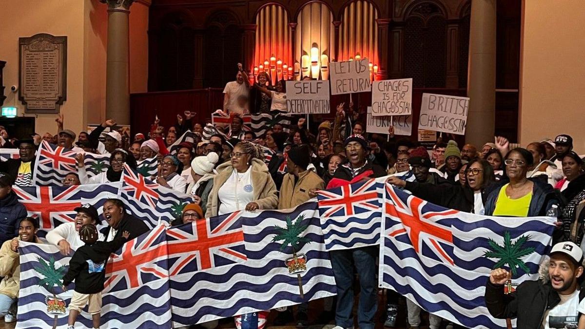 A group of about 100 people gathered in a hall with the people at the front of the group holding large flags of the Chagossians. They are standing in front of a large pipe organ.