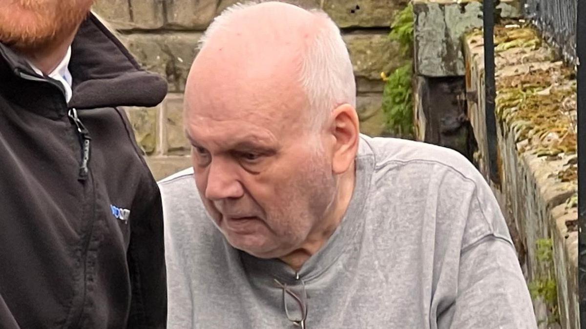 Richard Burrows, wearing a grey tracksuit with a pair of glasses tucked into the collar, looks away from the camera as he stands near a stone wall and next to a custody officer