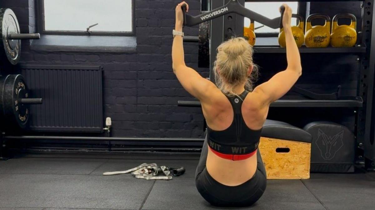 Victoria, working out in a gym, with her back to the camera. She has her arms up on a piece of machinery. She has her blonde hair tied up. Weights are to her left.