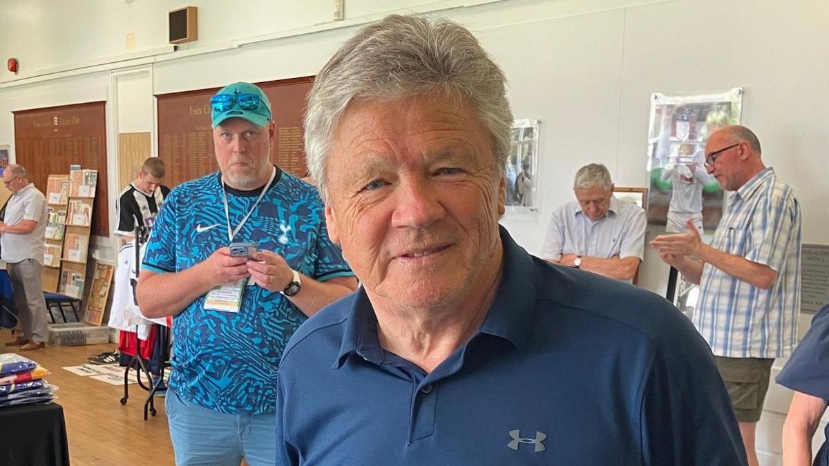 Steve Perryman smiles at the camera as if about to speak. He wears a navy blue jersey shirt with an open collar and one button done up He is clean shaven and has grey hair. Behind him a man in a blue baseball cap with a lanyard round his neck also looks at the camera. In the background, men mill about the stalls.