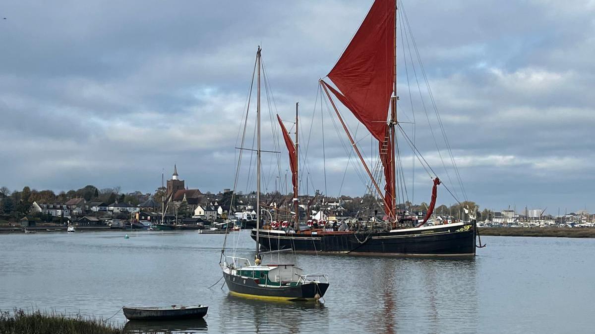 Two boats in frame, the one in the background significantly larger with a big red sail, water in the bottom half of the picture with a cluster of houses behind it on the top half