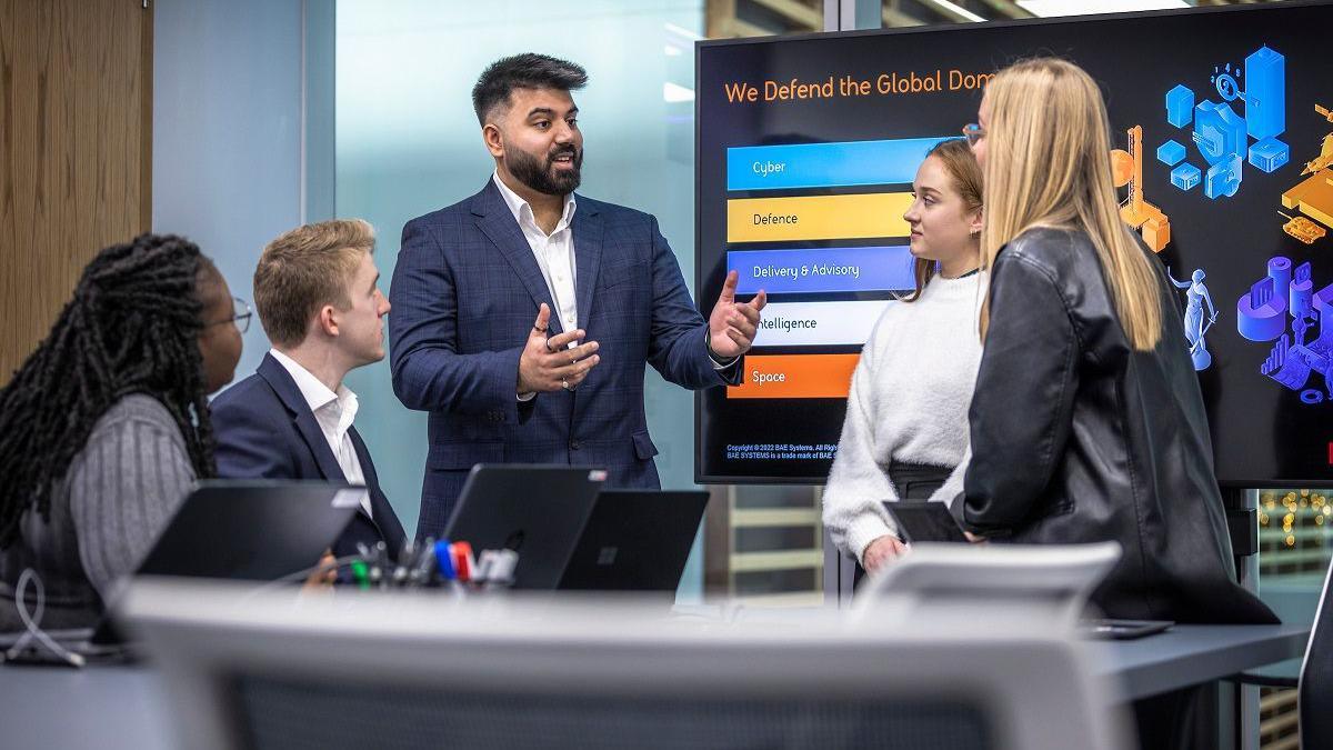 Two men and three women suits having a discussion in what looks like a company boardroom with a large screen behind them.