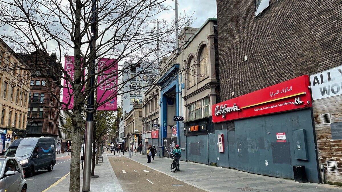 A view down Sauchiehall Street. It includes the road, cycle path and pedestrians on the pavement. Shops are boarded up on the right side of the street near the O2 ABC.