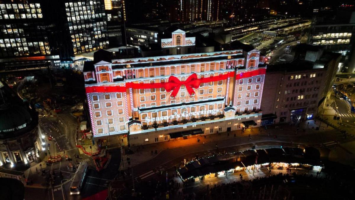 A large image of a gingerbread house with a red ribbon tied around it is projected onto the front of The Queen's Hotel in Leeds in 2023