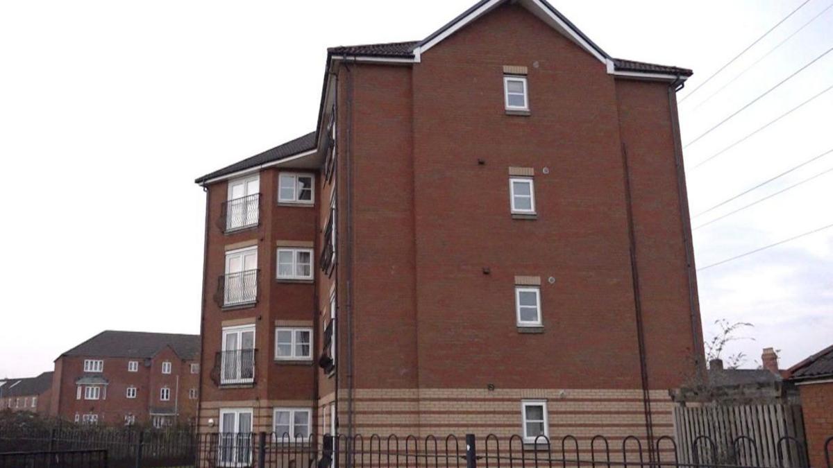 An orange brick block of flats with white windows and doors, surrounded by a black metal fence