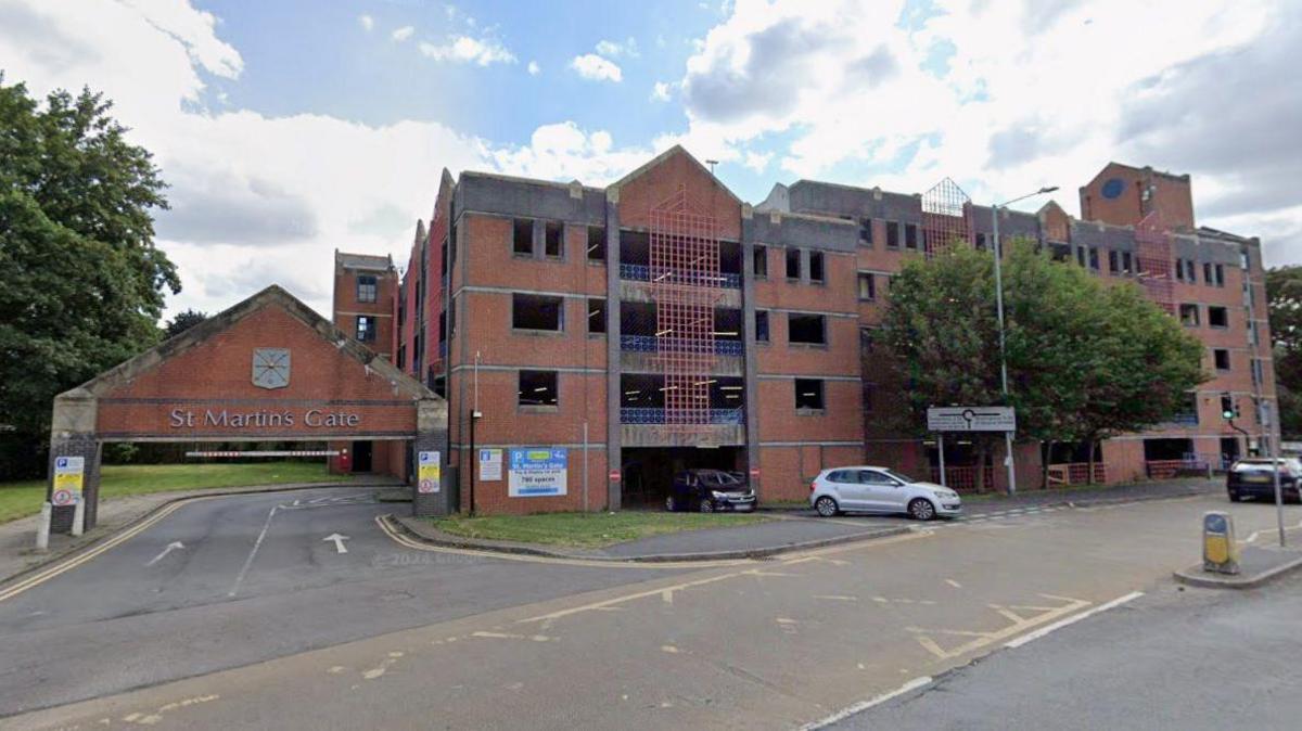 A street view image of a brick-built multi-storey car park. On the left is an entrance with a brick sign overhead that says "St Martin's Gate". Two cars can be seen coming out of the car park's exit on the right.