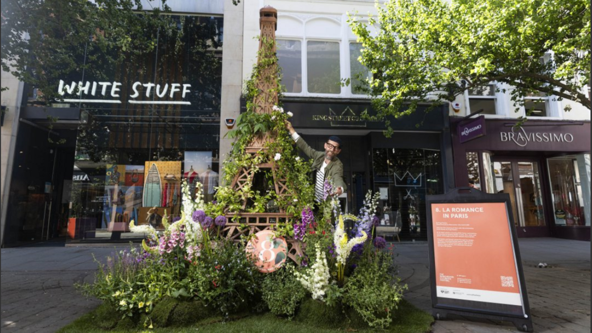 A floral Eiffel Tower surrounded by green shrubs and white and pink flowers. The artist stands to one side of it adjusting flowers. Behind him is a row of shops.