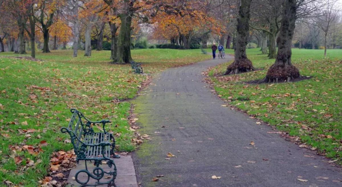 A gravel path winds through a park past a green metal bench. There are fallen leaves on the ground and several trees. In the distance, two men - one of them pushing a bike - are walking along the path.