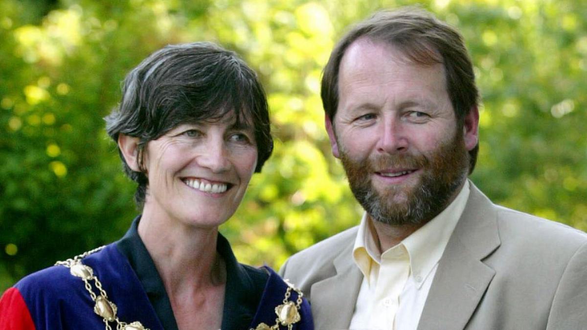 Catherine Connolly wearing her mayoral chains of office, poses for a photo with her husband Brian McEnery. She has short black hair and is smiling.   Her husband has dark, reddish hair and a beard and he is wearing a beige suit jacket and an open-neck yellow shirt.