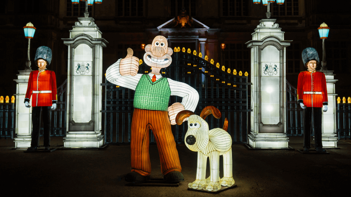 Characters Wallace, in a green jumper and brown trousers and wearing a tie, and Gromit, a dog looking up at Wallace, in front of Buckingham Palace-style gates and two Beefeaters standing to attention.
