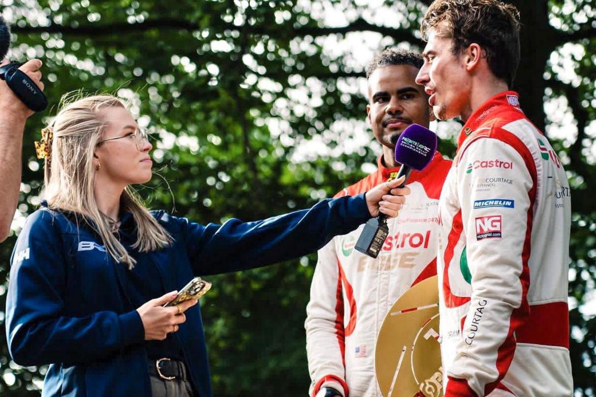 Hollie with blonde hair, wearing glasses, a dark blue jacket, and olive-green pants is holding a microphone towards a man on the right. She has a phone in her other hand. The man on the right is wearing a white and red racing suit with green accents. He is looking at the reporter and appears to be speaking. Behind him stands another man in a similar racing suit, holding a golden trophy.