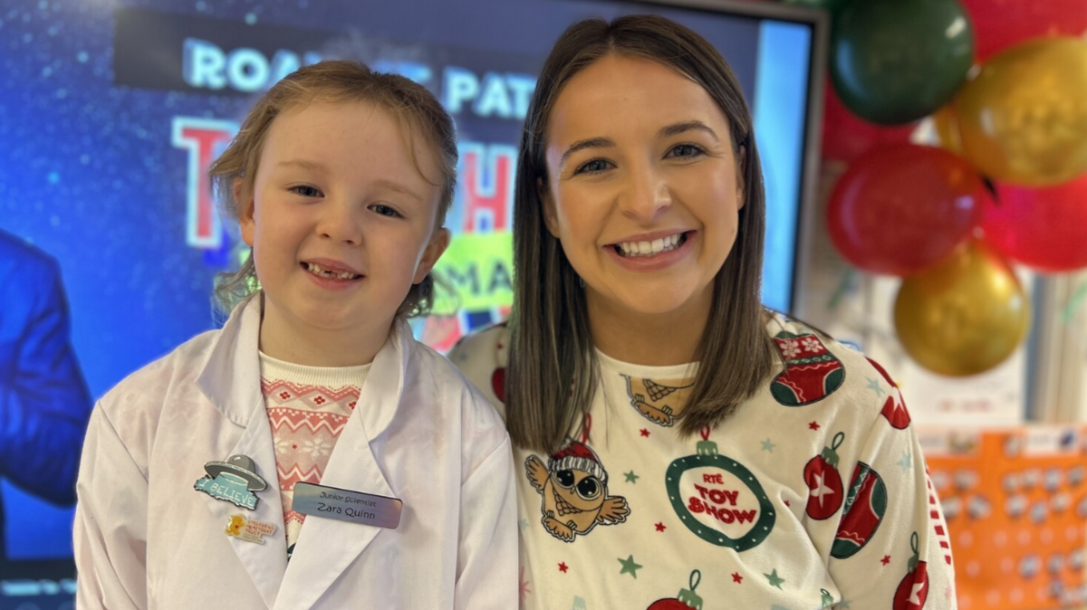 Zara is dressed in a white scientist coat, and standing beside her teacher who is wearing pyjamas