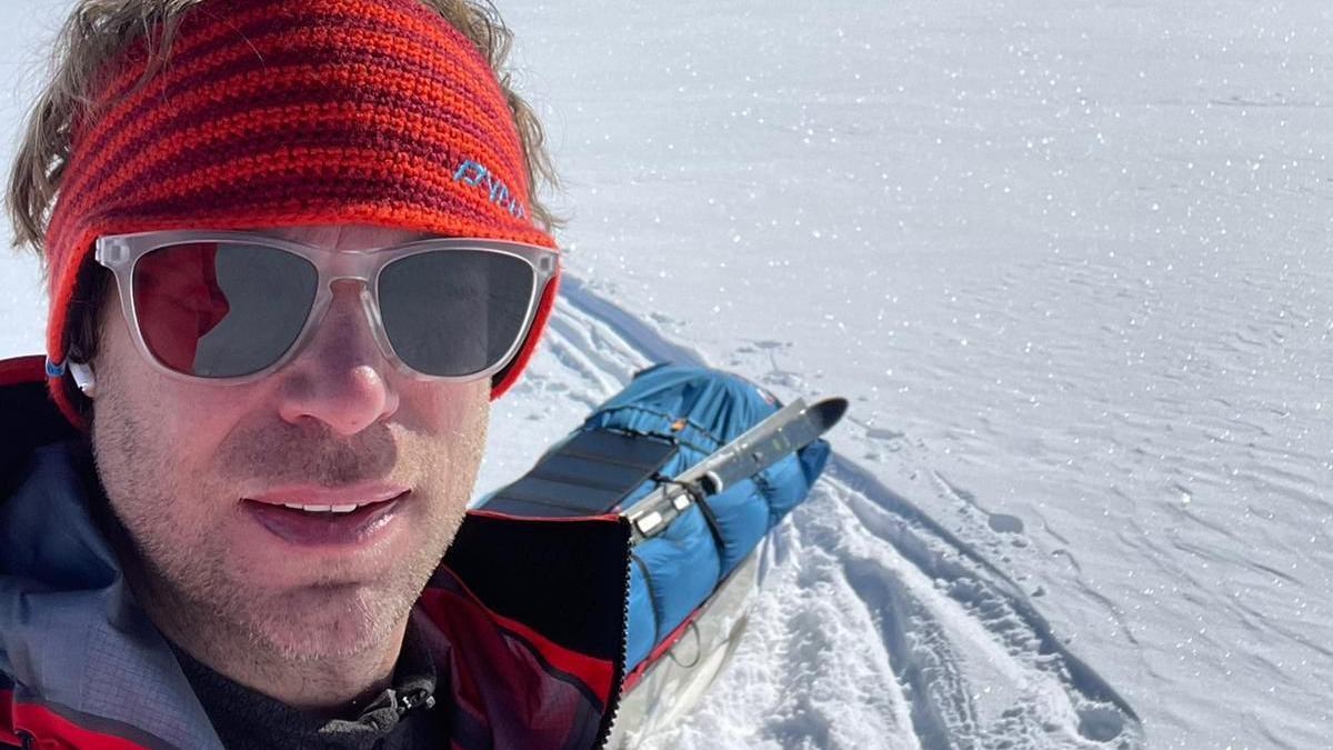 A selfie of Jonny Huntington with his kit on the snow behind him. He is wearing sunglasses and a red knitted headband and a red coat.