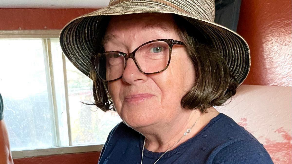 A woman wearing a straw sun hat and glasses smiles at the camera. She has brown hair in a bob, and is wearing a navy blue top and a silver necklace. In the background is a wall painted brick red and a window.