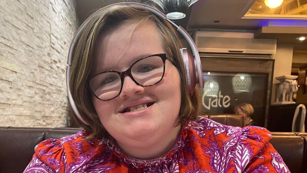 Phoebe Ockenden wearing a red floral dress and bluetooth headphones, sitting in a restaurant.