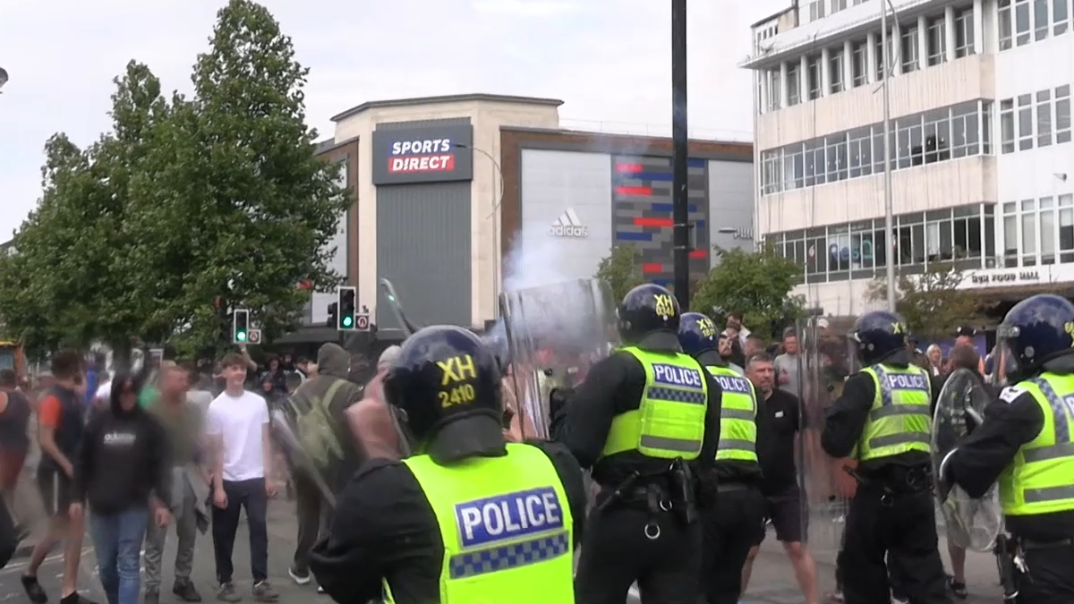 Police officers in riot gear face rioters during the unrest that followed the stabbing of three children in Southport last summer. The police officers are holding large clear shields, facing down would-be rioters. A Sports Direct store stands on a street corner in the background.