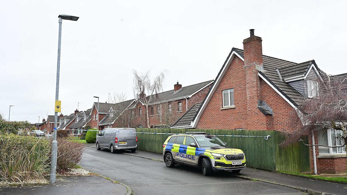 A yellow and blue police car sits behind blue and white police tape. It is mounted on the black footpath in-front of a collection of red brick houses