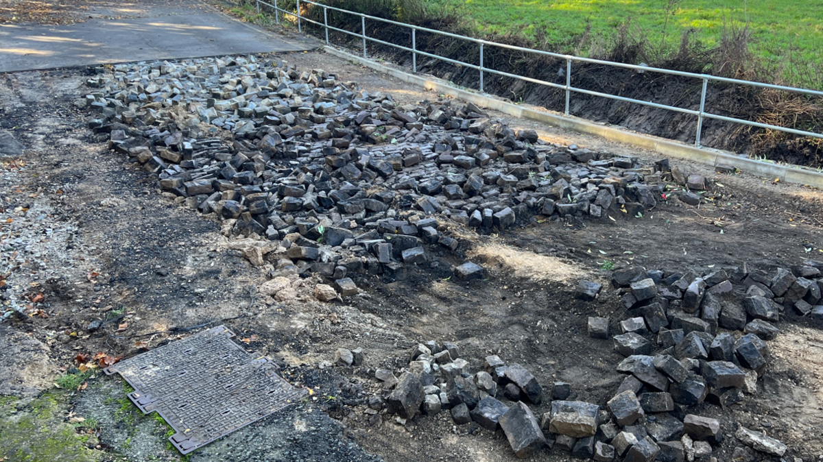 Image shows Mill Lane road and chunks of cobbles which have been dug up. They are in a large pile in the centre of the road where a rectangle section has been ripped away by flooding. To the far right of the image is a large green field. 