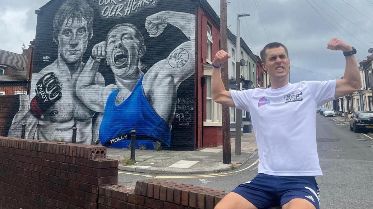Joe Wild during his marathon in Liverpool, posing with his biceps flexed in front of a mural of martial arts fighters Paddy Pimblett and Molly McCann.