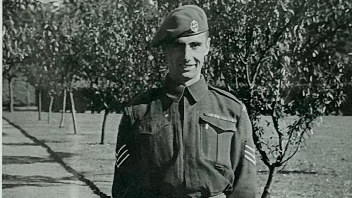 Black and white historical image of a man in Army uniform with a beret on. He is stood outdoors with a row of trees in leaf behind him.