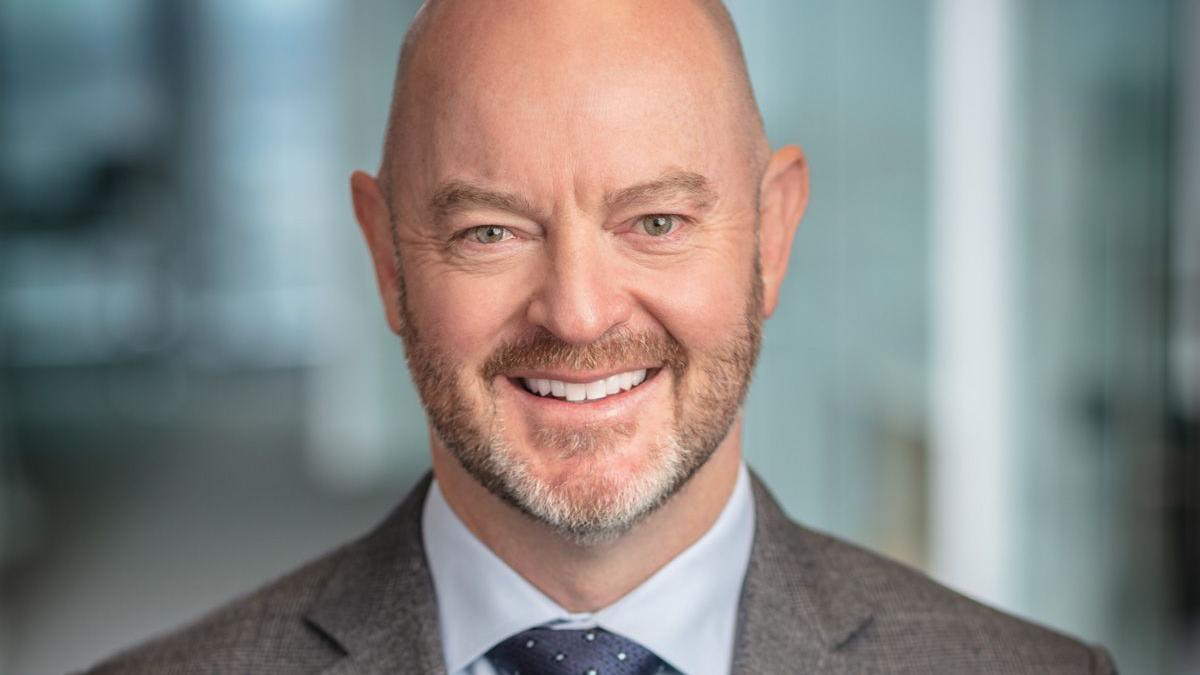 A professional headshot of Leigh Curyer smiling, wearing a grey suit jacket, a light blue button-up shirt and a speckled dark blue tie.