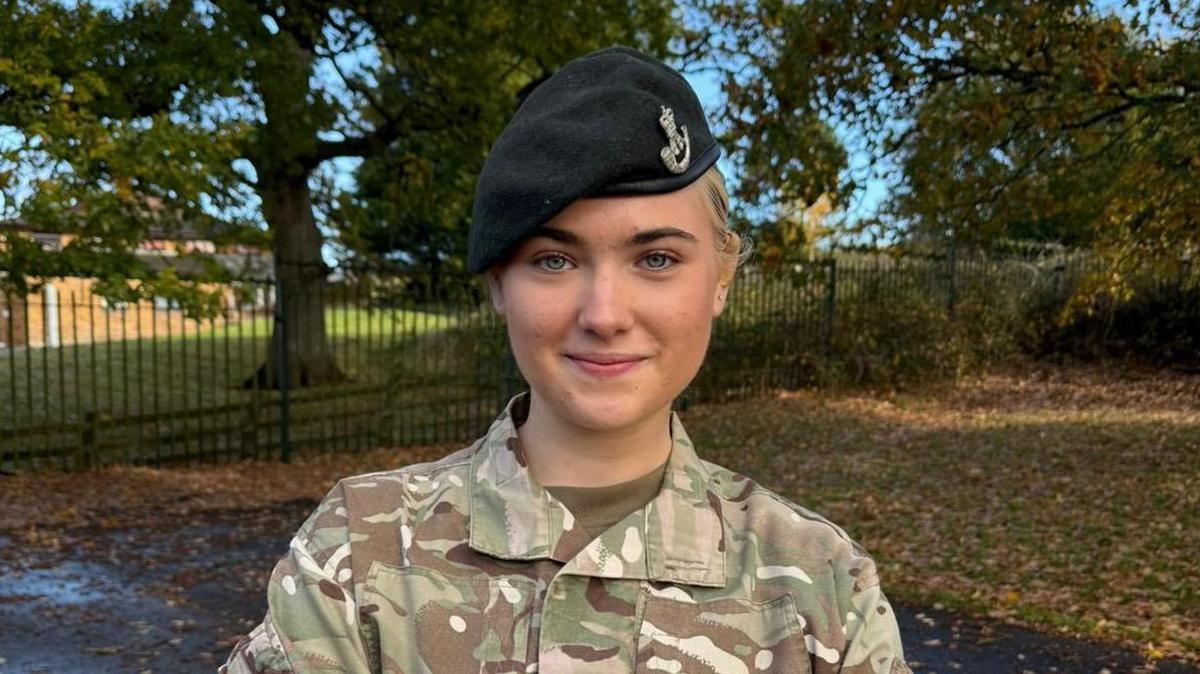 Arabella Mason pictured standing in a park on an autumnal day. She has blonde hair which is tied back in a ponytail and is wearing her Army Cadet Force camouflage uniform and hat.
