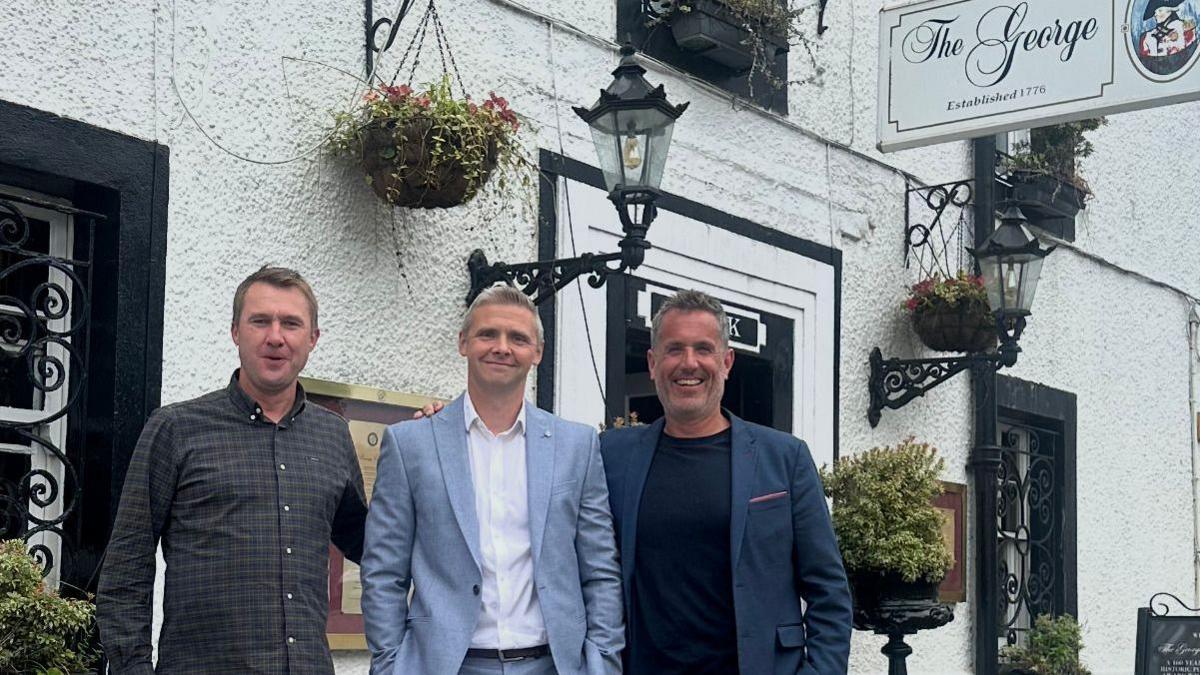 Three men standing outside a hotel - they are all smiling and grinning.