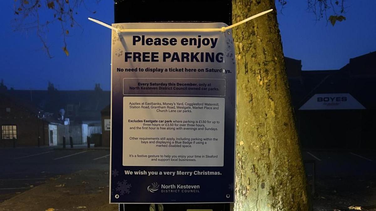 A sign attached to a parking meter at night, offering free parking on Saturdays in December in North Kesteven District Council-owned car parks. A tree and buildings stand in the background.