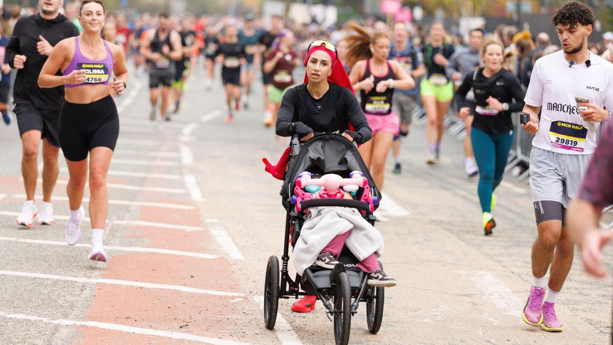 Aneila Afsar, who is wearing a dark outfit and red hijab scarf, runs while pushing a buggy with her child. She is surrounded by scores of other participants as they run down a wide road.