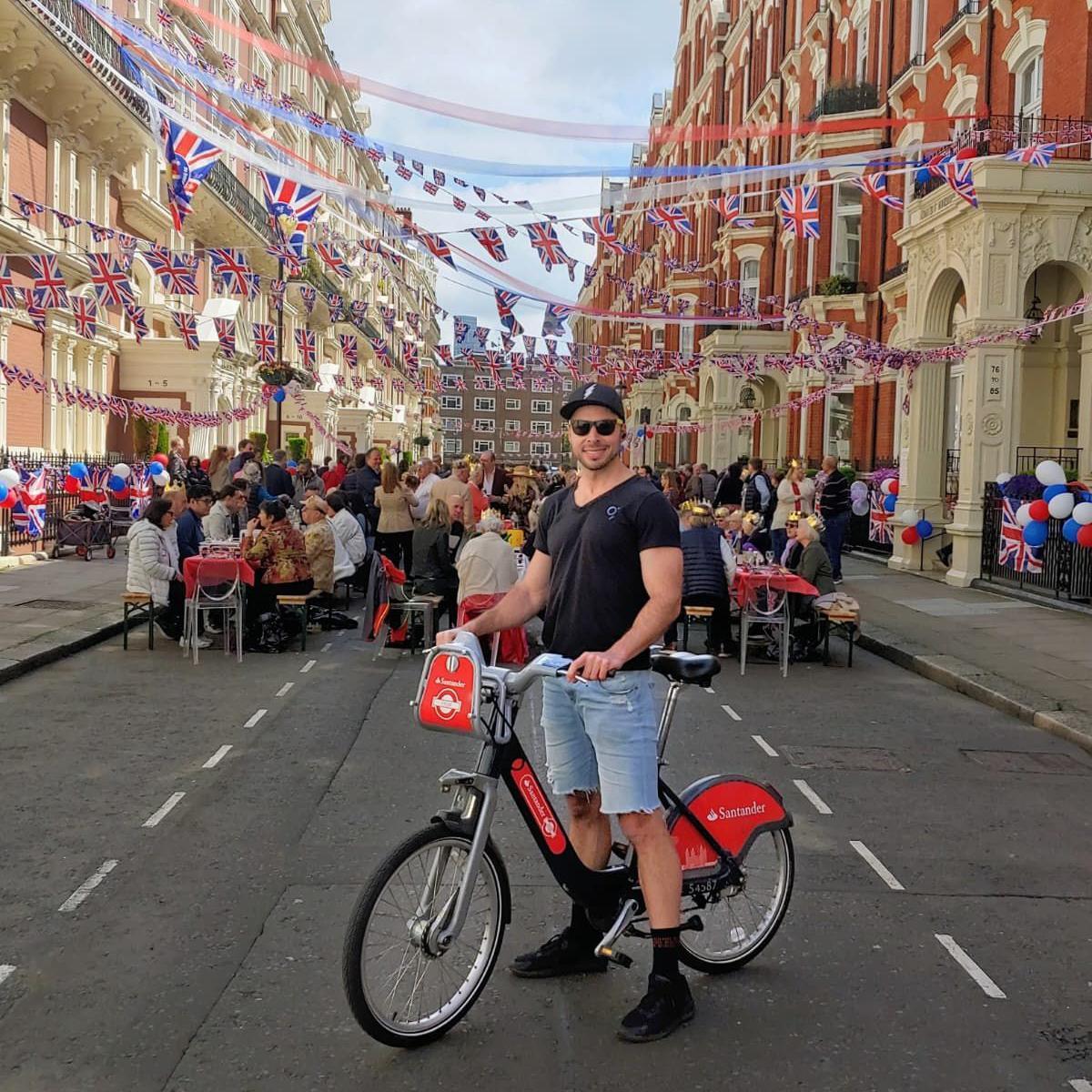 A man wearing sunglasses, a black T-shirt and denim shorts stands with a Santander Cycle on a street decorated with Union Jack bunting. Behind him, long tables are set up in the road with people gathered for a coronation street party.
