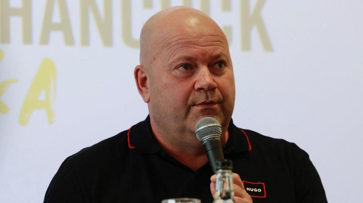 Mark Porter is holding a microphone in front of a mainly white background. He is wearing a dark top.