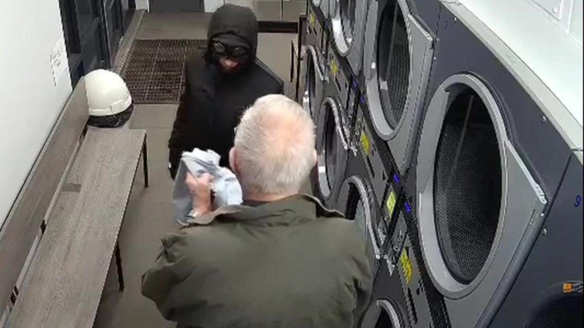 A man with his back to camera holding a pair of jeans faces a man in black jacket and black mask. They are inside a launderette.