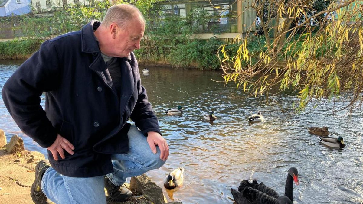 Anthony James, dressed in a black coat and blue jeans, looking at a canal that has a black swan and ducks.