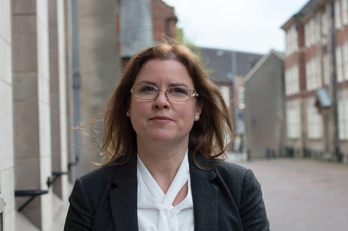 Victoria Wilson looks into the camera. She wears rimless glasses and has dark brown hair to her shoulders. She wears a dark jacket over a white top and stands in a thoroughfare between buildings which are blurred behind her.