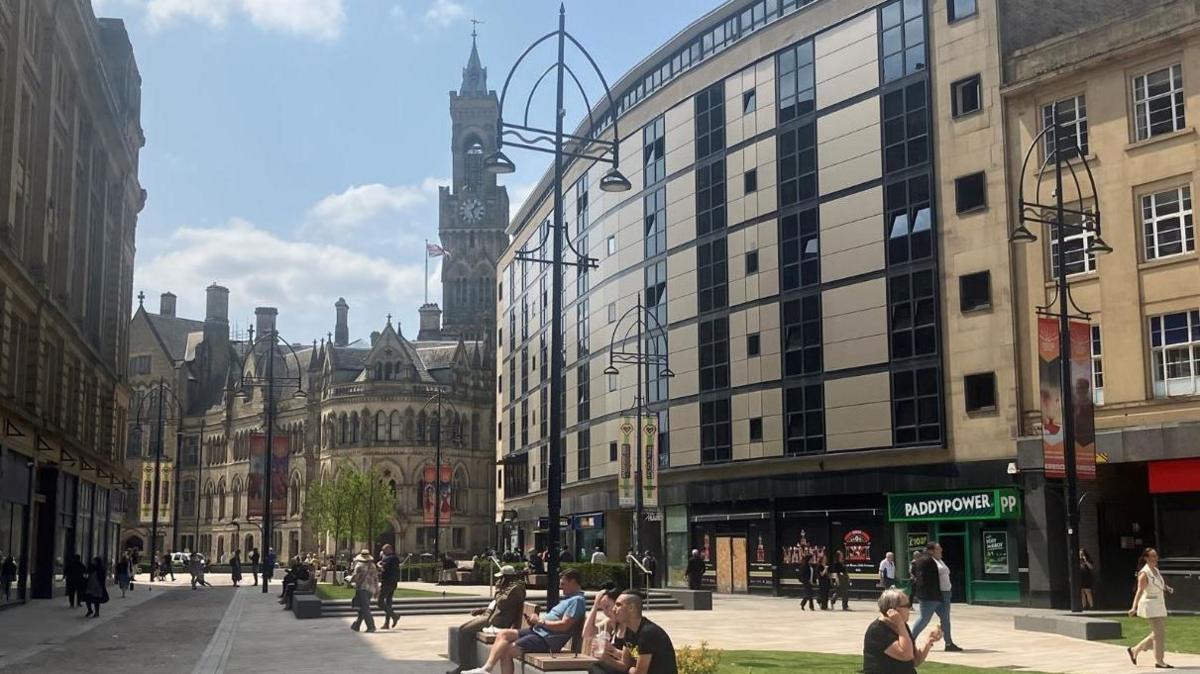 People sit on benches in a pedestrianised area with historical buildings in the background and more modern ones flanking the paved area.