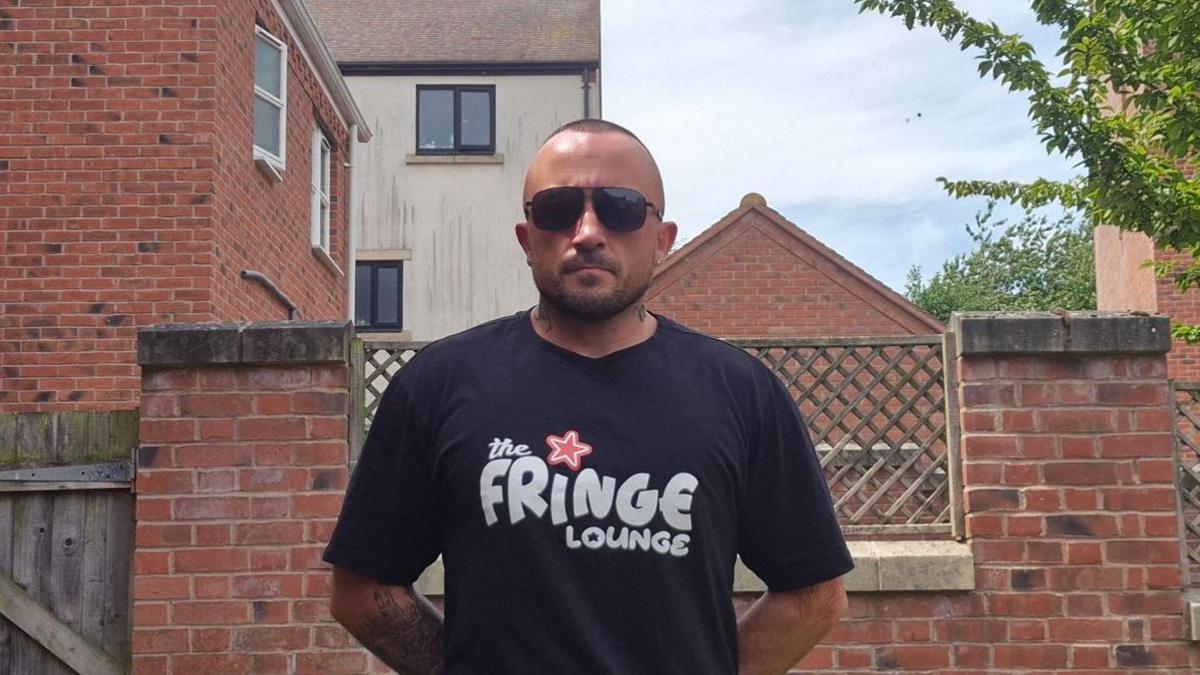 A man in dark glasses and a dark tshirt with the words the fringe lounge on it, standing in front of a brick wall