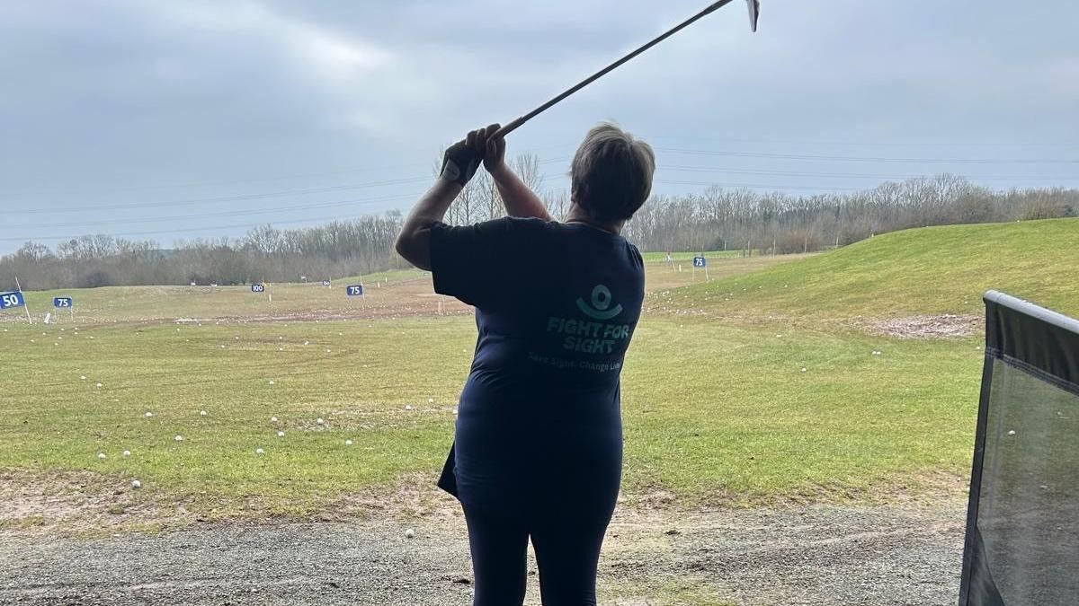 An action shot of a woman swinging a golf club on a driving range. The sky is cloudy in the distance. She has the golf club above her head after hitting the ball. The woman is wearing a blue top and leggings. The top has a logo on it that reads "Fight for Sight".