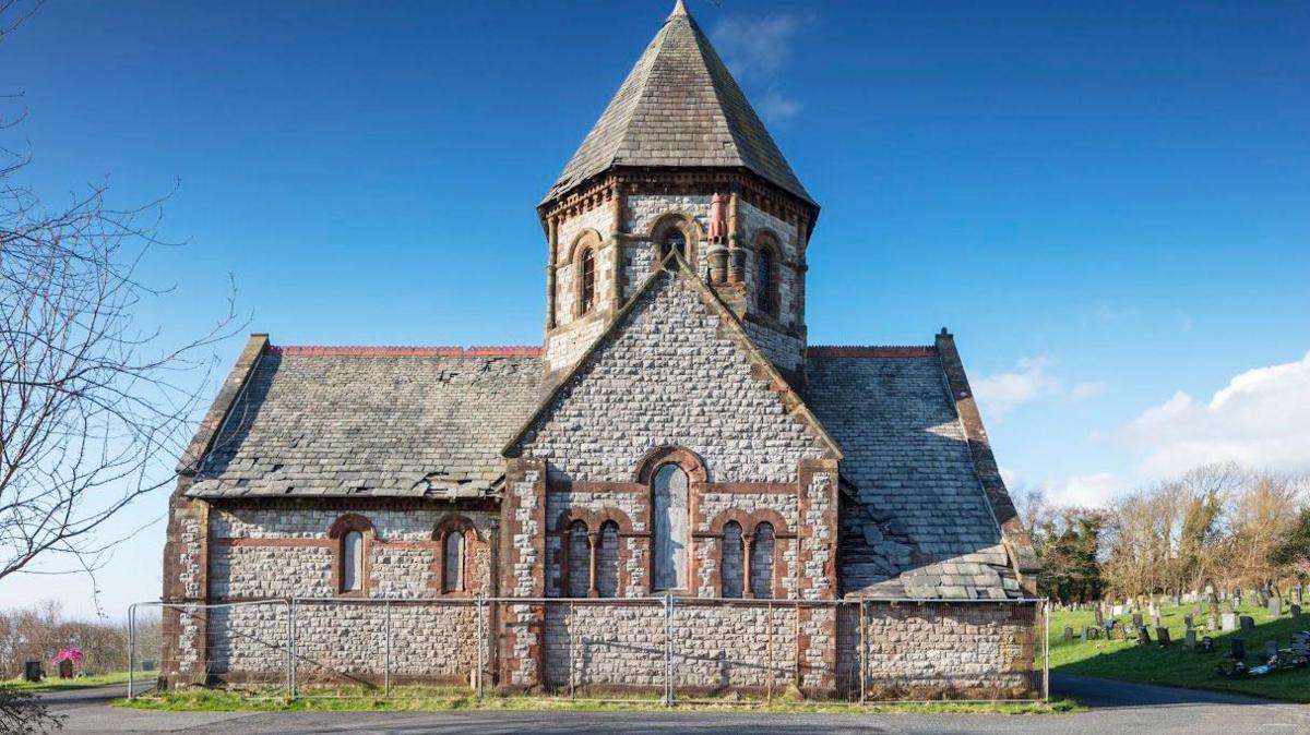 A chapel in Barrow Cemetery on a sunny day. The building has windows but they have been boarded up. The building is Victorian-style with large white and red bricks. 